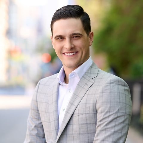 Professional headshot of a smiling man in a light gray blazer and lavender shirt against a blurred urban background